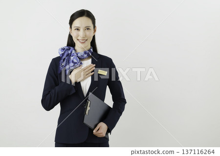 A smiling Japanese woman concierge wearing a navy suit with a ribbon and name tag on a white background, holding her hand to her chest A smiling Japanese woman concierge wearing a navy suit with a ribbon and name tag on a white background, holding her hand to her chest 137116264