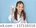A funny girl in a white coat in the guise of a pediatrician holds a stethoscope. Pediatric cardiology, diseases of the heart and cardiovascular system 137116318