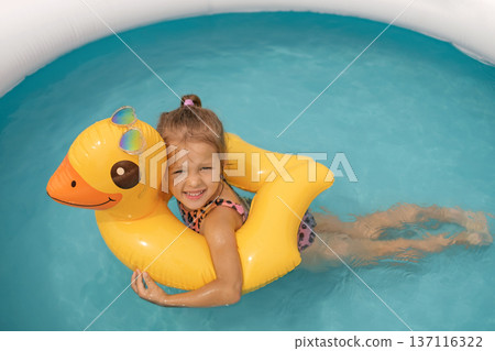 A little girl is happily swimming in a small inflatable pool, hugging a duck-shaped swimming circle. Summer vacation vacation. Skin damage caused by sunburn 137116322