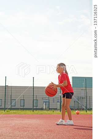 A little girl holds a basketball in hands, gathering her strength before throwing.  A popular toy for marksmanship training 137116325