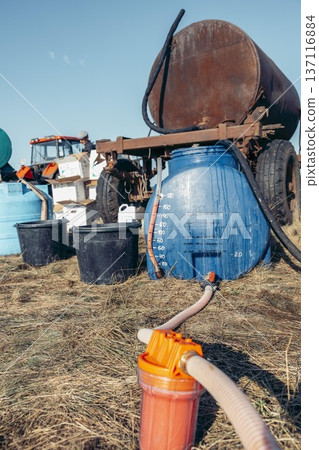 Farm equipment for chemical spraying. A large blue tank and a rusty trailer are visible. Buckets are placed nearby on dry grass under a clear sky. 137116884