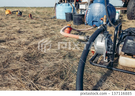 Farm equipment with a pump and hoses on dry grass. Blue tanks are visible in the background, indicating agricultural chemical spraying activities. Farm equipment with a pump and hoses on dry grass. Blue tanks are visible in the background, indicating agricultural chemical spraying activities. 137116885