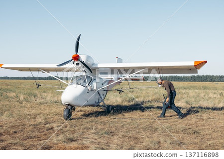 man in protective mask go to refueling small white aircraft on grassy airfield, aviation worker in green uniform during sunset, rural field, aviation service, pilot training, countryside tourism 137116898