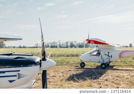 Two airplanes parked on a farm field. One is a small white aircraft with a red stripe, and the other is a black and white plane. The background features green fields under a clear sky. Two airplanes parked on a farm field. One is a small white aircraft with a red stripe, and the other is a black and white plane. The background features green fields under a clear sky. 137116914
