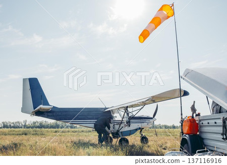 man refueling small blue aircraft on grassy airfield, aviation worker in green uniform during sunset, rural field, aviation service, pilot training, countryside tourism 137116916