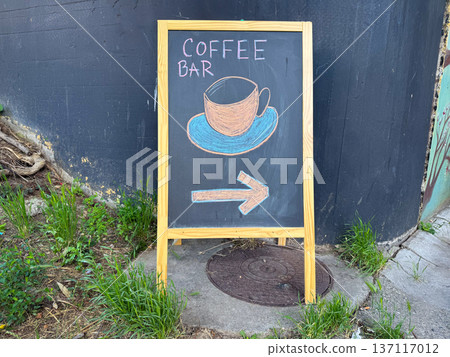 Chalkboard sign with drawing of coffee cup and arrow standing on street near wall and grass outdoors. Cafe direction sign, street advertising, urban cafe culture and small business navigation Chalkboard sign with drawing of coffee cup and arrow standing on street near wall and grass outdoors. Cafe direction sign, street advertising, urban cafe culture and small business navigation 137117012
