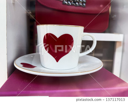White coffee cup with red heart symbol placed on saucer in shop window display with pink handbag in background. Romantic gift idea, love symbol, valentine mood and cafe lifestyle aesthetic White coffee cup with red heart symbol placed on saucer in shop window display with pink handbag in background. Romantic gift idea, love symbol, valentine mood and cafe lifestyle aesthetic 137117013