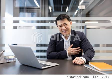 Asian businessman experiencing a sudden heart attack or acute chest pain while working at his office desk, clutching his chest in discomfort and showing an expression of intense physical suffering Asian businessman experiencing a sudden heart attack or acute chest pain while working at his office desk, clutching his chest in discomfort and showing an expression of intense physical suffering 137117017