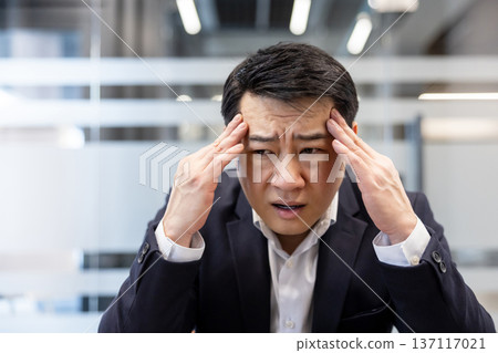 Asian businessman experiencing a severe headache and high stress while working in a modern office, pressing temples to relieve the pain and showing a frustrated expression Asian businessman experiencing a severe headache and high stress while working in a modern office, pressing temples to relieve the pain and showing a frustrated expression 137117021