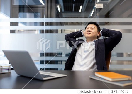 Asian businessman reclining in office chair with hands behind head, eyes closed, smiling contentedly at a laptop on the desk after finishing work and enjoying a calm moment of success Asian businessman reclining in office chair with hands behind head, eyes closed, smiling contentedly at a laptop on the desk after finishing work and enjoying a calm moment of success 137117022