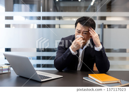Asian businessman working in a modern office, experiencing flu symptoms and a headache, blowing his nose with a tissue while focused on his laptop, depicting illness and discomfort at work Asian businessman working in a modern office, experiencing flu symptoms and a headache, blowing his nose with a tissue while focused on his laptop, depicting illness and discomfort at work 137117023