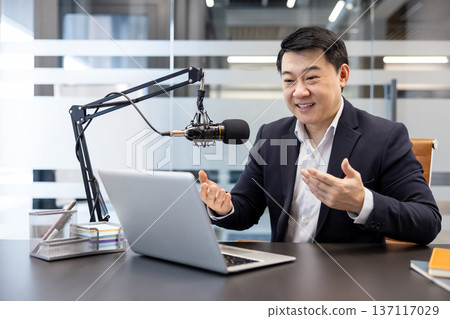 Asian man speaking into a professional microphone and looking at a laptop, broadcasting content in a modern office studio, sharing communication and creative ideas Asian man speaking into a professional microphone and looking at a laptop, broadcasting content in a modern office studio, sharing communication and creative ideas 137117029
