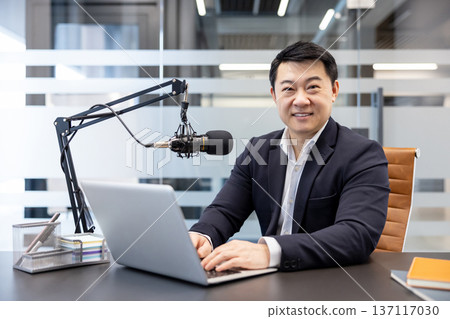 Asian man smiling at camera while working on laptop and recording a podcast or broadcasting from a modern office studio, setup with a professional microphone on his desk Asian man smiling at camera while working on laptop and recording a podcast or broadcasting from a modern office studio, setup with a professional microphone on his desk 137117030