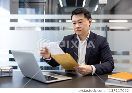Serious asian businessman holding and opening a large mail envelope with official documents or a letter while sitting at his desk with a laptop, managing communication and work 137117032
