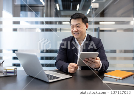 Asian businessman smiling and focused, working productively using a laptop and a digital tablet, sitting at a desk in a modern corporate office environment, representing success and professionalism 137117033