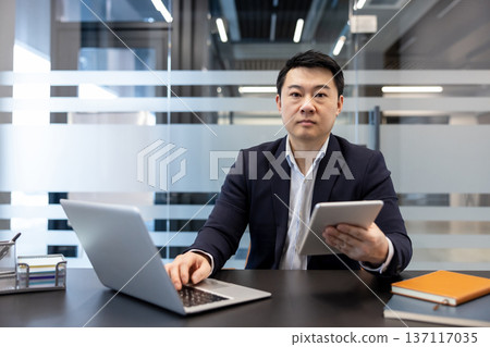Young asian businessman in suit multitasking at office desk, looking at camera while using laptop and holding a tablet, focused on digital workflow and productivity Young asian businessman in suit multitasking at office desk, looking at camera while using laptop and holding a tablet, focused on digital workflow and productivity 137117035