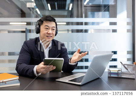 Asian businessman wearing headphones participating in an online video call meeting, talking and gesturing while holding a notebook, working remotely from a modern office 137117056