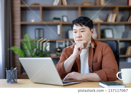 Asian man sitting at a desk, looking at his laptop screen with a pensive expression, representing concentration, remote work, and focused digital activity from a comfortable home office setting Asian man sitting at a desk, looking at his laptop screen with a pensive expression, representing concentration, remote work, and focused digital activity from a comfortable home office setting 137117067