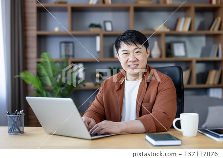 Smiling asian man in smart-casual attire types on a laptop at a wooden desk in a modern home office, looking at the camera with confident, relaxed professionalism Smiling asian man in smart-casual attire types on a laptop at a wooden desk in a modern home office, looking at the camera with confident, relaxed professionalism 137117076