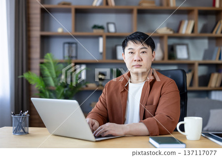 Asian man sitting at a desk, looking at the camera while typing on a laptop, surrounded by office elements, symbolizing productivity, focus, and remote work in a comfortable environment 137117077