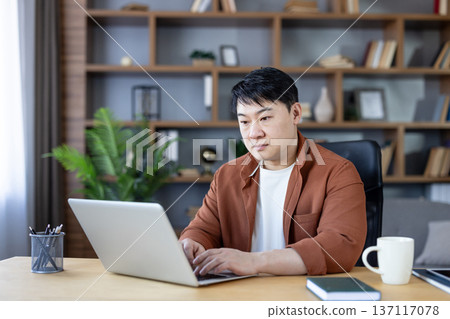 Asian man focused on laptop in a modern home office, working remotely on freelance business tasks at his desk, typing, researching and planning a project Asian man focused on laptop in a modern home office, working remotely on freelance business tasks at his desk, typing, researching and planning a project 137117078