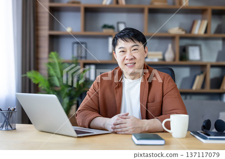Asian man sitting at desk with laptop, smiling confidently and looking at camera, representing remote work, freelance entrepreneurship, and productivity in a modern home office setting Asian man sitting at desk with laptop, smiling confidently and looking at camera, representing remote work, freelance entrepreneurship, and productivity in a modern home office setting 137117079