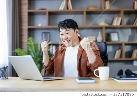 Asian man in home office celebrating success with clenched fists and broad smile while looking at laptop, joyful freelancer reacting to good news or winning message Asian man in home office celebrating success with clenched fists and broad smile while looking at laptop, joyful freelancer reacting to good news or winning message 137117080