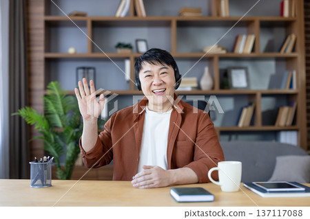 Smiling asian man wearing a headset and brown shirt, sitting at a desk in a home office, looking at the camera and waving during a virtual meeting, video conference, or online communication Smiling asian man wearing a headset and brown shirt, sitting at a desk in a home office, looking at the camera and waving during a virtual meeting, video conference, or online communication 137117088