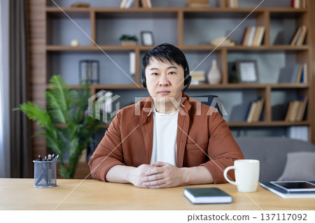 Asian man in a home office wearing a headset, focused on a video call or webinar with hands clasped on the desk beside laptop, notebook and mug Asian man in a home office wearing a headset, focused on a video call or webinar with hands clasped on the desk beside laptop, notebook and mug 137117092