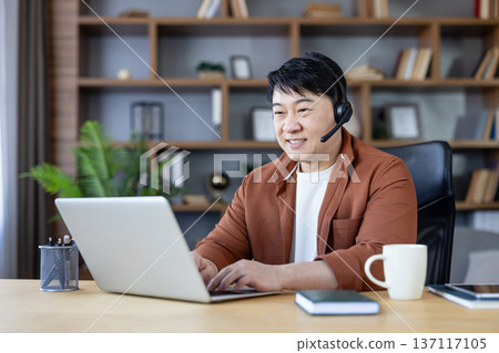 Asian man working from home office, typing on a laptop and wearing a headset, engaging in a video call or online meeting, creating a productive and focused work environment 137117105