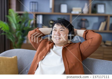 Asian man smiling and relaxing on a comfortable couch in a living room, enjoying leisure time and listening to music through black headphones, finding peace and tranquility at home Asian man smiling and relaxing on a comfortable couch in a living room, enjoying leisure time and listening to music through black headphones, finding peace and tranquility at home 137117111