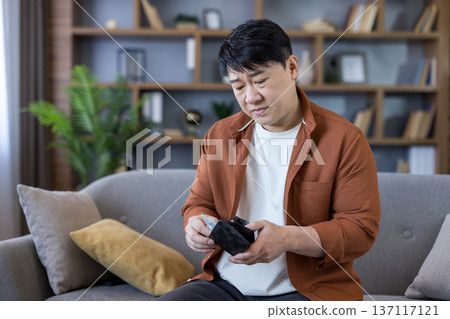 Asian man sitting on a sofa, looking worried while checking his almost empty wallet, indicating financial problems, budget constraints, and economic struggles 137117121