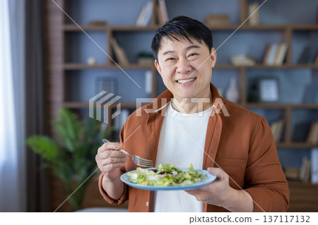 Smiling asian man at home holding a plate of fresh green salad and fork, enjoying a healthy, balanced meal and promoting wellness, mindful eating and contentment 137117132