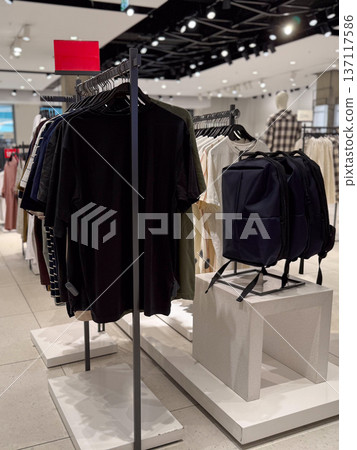 Clothing and backpacks on racks are displayed in a bright modern retail store interior. Consumer culture, commerce, lifestyle and seasonal shopping trends in contemporary fashion industry. Clothing and backpacks on racks are displayed in a bright modern retail store interior. Consumer culture, commerce, lifestyle and seasonal shopping trends in contemporary fashion industry. 137117586