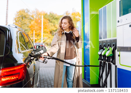 Beautiful woman filling car with gasoline at gas station. Online pay. Woman using app on phone. Beautiful woman filling car with gasoline at gas station. Online pay. Woman using app on phone. 137118433