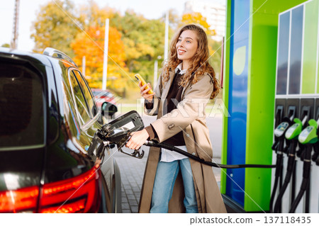 Beautiful woman filling car with gasoline at gas station. Online pay. Woman using app on phone. Beautiful woman filling car with gasoline at gas station. Online pay. Woman using app on phone. 137118435