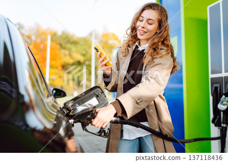 Beautiful woman filling car with gasoline at gas station. Online pay. Woman using app on phone. 137118436