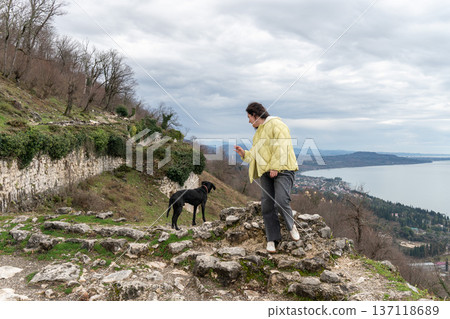Woman stepping on stone ruins near black dog overlooking New Athos coastline and Black Sea from Anacopia fortress in Abkhazia under cloudy sky, Travel lifestyle scene suitable for tourism marketing Woman stepping on stone ruins near black dog overlooking New Athos coastline and Black Sea from Anacopia fortress in Abkhazia under cloudy sky, Travel lifestyle scene suitable for tourism marketing 137118689
