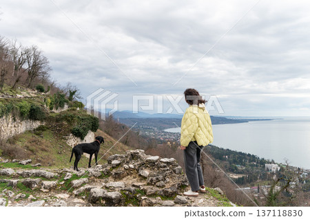 Woman and black dog standing on Anacopia fortress ruins overlooking New Athos coastline and Black Sea in Abkhazia under cloudy sky, Outdoor travel scene suitable for tourism marketing Woman and black dog standing on Anacopia fortress ruins overlooking New Athos coastline and Black Sea in Abkhazia under cloudy sky, Outdoor travel scene suitable for tourism marketing 137118830