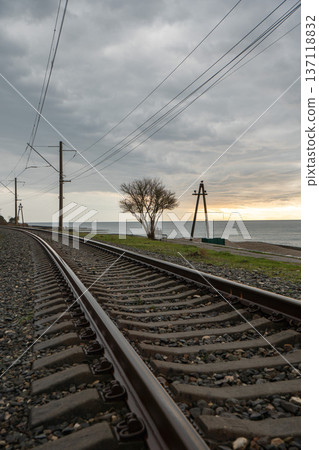 Railway tracks along Black Sea coastline in Abkhazia under cloudy sky with power lines and lone tree near shore, Transport infrastructure landscape suitable for travel marketing, regional promotion Railway tracks along Black Sea coastline in Abkhazia under cloudy sky with power lines and lone tree near shore, Transport infrastructure landscape suitable for travel marketing, regional promotion 137118832