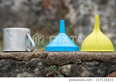 Metal mug, plastic funnels in blue, yellow standing on stone surface outdoors, Household utensils, simple objects suitable for product presentation, minimal lifestyle and everyday utility concepts Metal mug, plastic funnels in blue, yellow standing on stone surface outdoors, Household utensils, simple objects suitable for product presentation, minimal lifestyle and everyday utility concepts 137118833