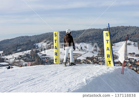 Rear view of a skier standing between yellow start flags atop a snowy slope. 137118838