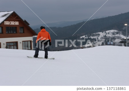 snowboarder gliding down a snowy mountain slope near a wooden chalet 137118861