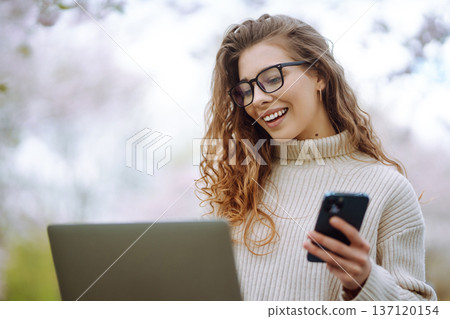Cheerful woman on phone takes selfie while sitting at table with laptop in spring park. Lifestyle. Cheerful woman on phone takes selfie while sitting at table with laptop in spring park. Lifestyle. 137120154