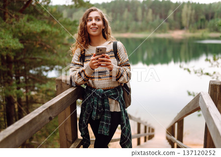 A young woman in a cozy sweater walking near a beautiful lake, smiling and using her phone A young woman in a cozy sweater walking near a beautiful lake, smiling and using her phone 137120252