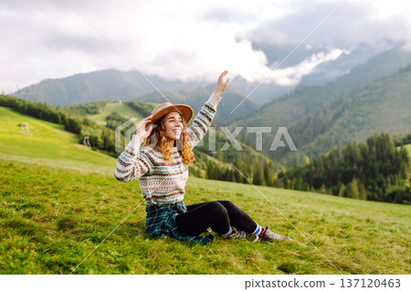 Stylish female traveler in hat has fun on top of hill, enjoys mountain landscape. Active lifestyle. 137120463