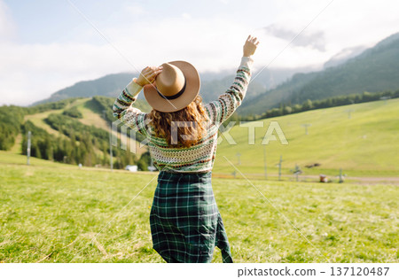 Stylish female traveler in hat has fun on top of hill, enjoys mountain landscape. Active lifestyle. 137120487