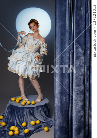 Young woman in vintage dress standing on pedestal with lemons in studio with blue curtain Young woman in vintage dress standing on pedestal with lemons in studio with blue curtain 137121012