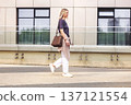 Stylish woman walking outdoors in modern city, wearing casual neutral outfit with gray t-shirt, white trousers and sneakers, beige striped shirt tied at waist , carrying brown handbag. Smiling female 137121554