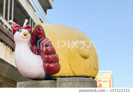 Playful snail sculpture with bow standing on concrete pedestal under blue sky. Whimsical street art, vivid decoration, joyful urban detail bringing charm and character to city space. Playful snail sculpture with bow standing on concrete pedestal under blue sky. Whimsical street art, vivid decoration, joyful urban detail bringing charm and character to city space. 137122151